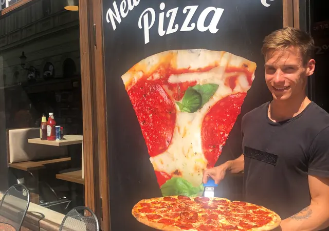 Muž drží celú pizzu so salámou pred výkladom s nápisom New York Style Pizza