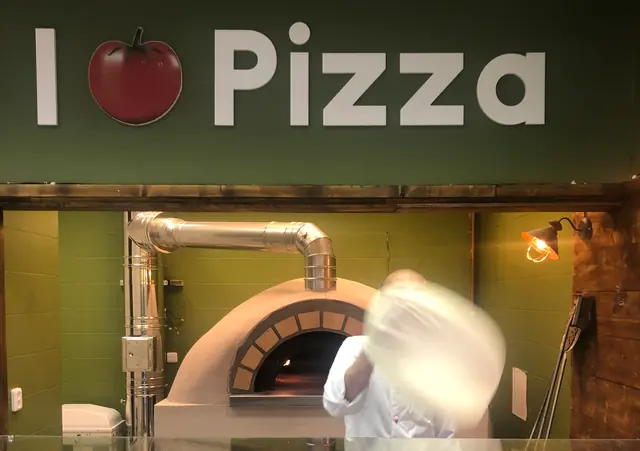 Pizzaiolo vyhadzuje cesto pred pecou pod nápisom I Love Pizza na zelenej stene