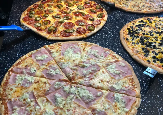 Viacero celých pizz na tmavom pulte, vpredu pizza so šunkou a syrom nakrájaná na kúsky